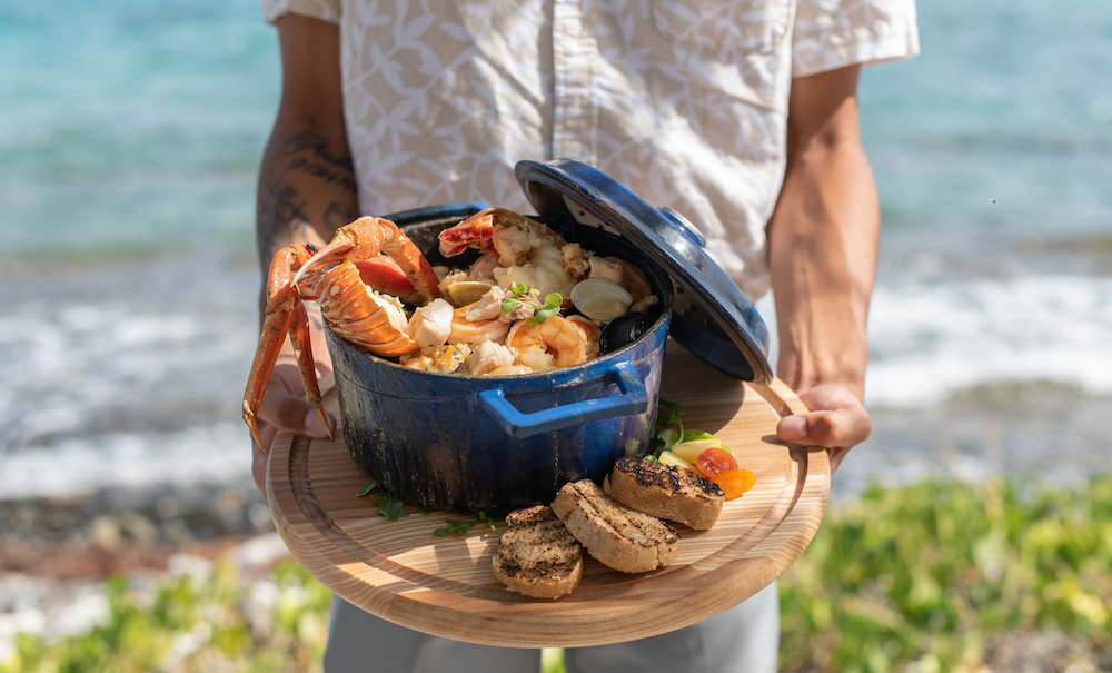 Person holding a pot of seafood and bread near the ocean.