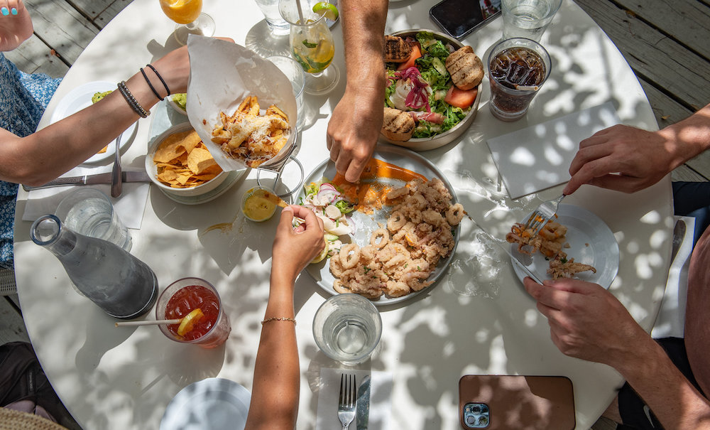 People sharing various dishes around a round outdoor table with drinks and utensils.