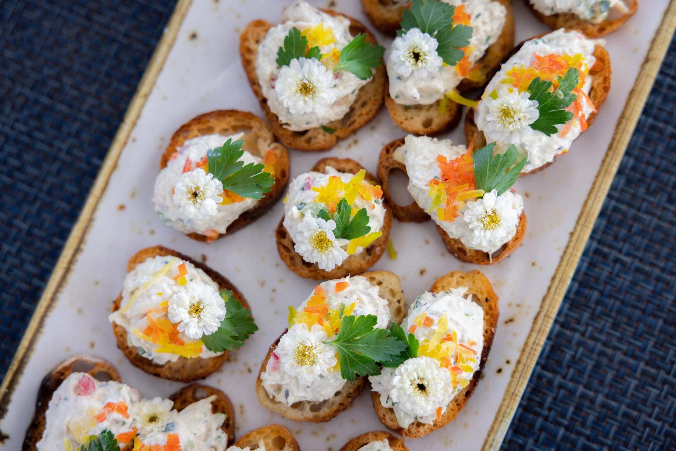 Tray of crostini topped with cream, herbs, edible flowers, and citrus zest on a dark surface.