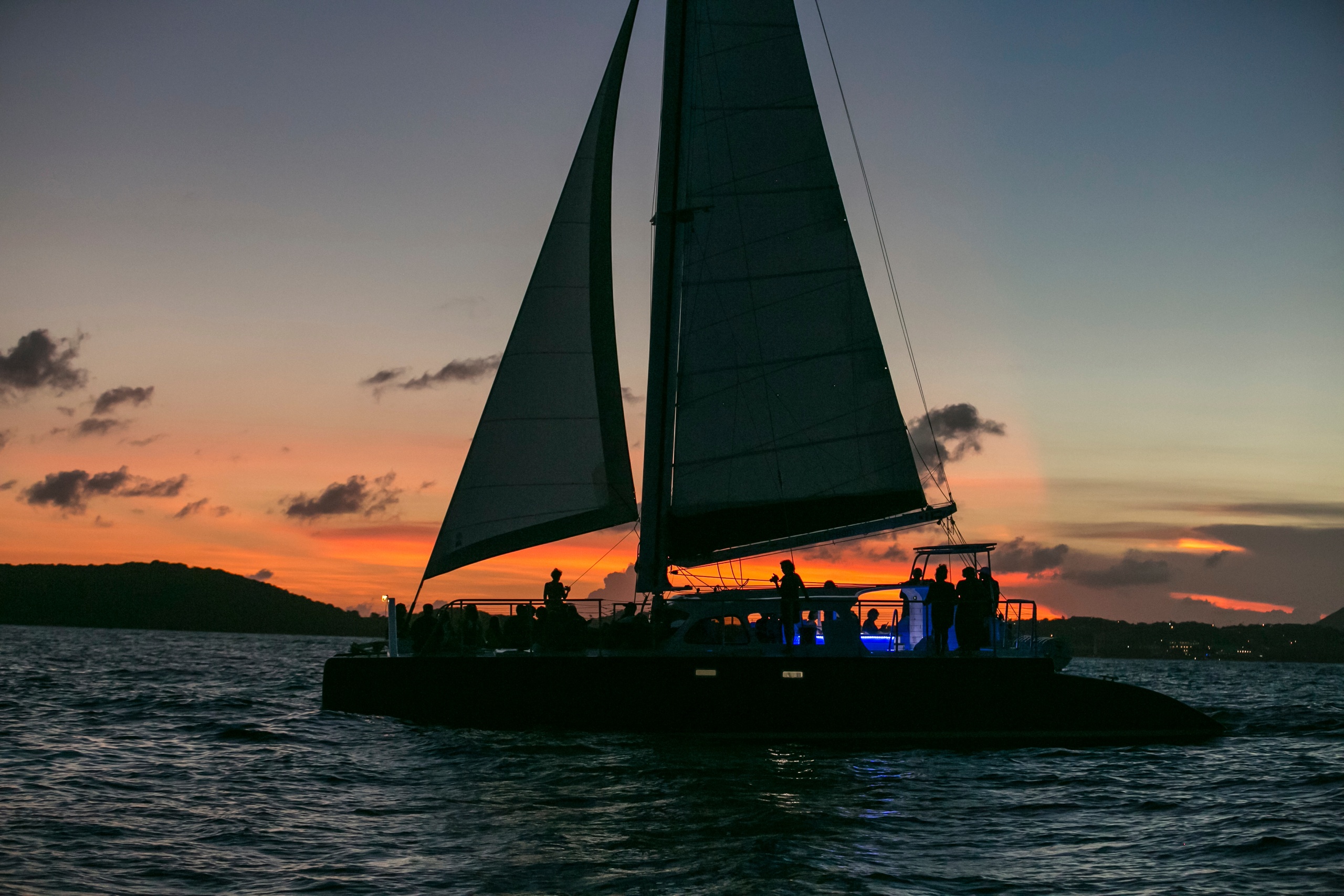 Sailboat on the water at sunset with dark silhouetted sails and orange sky.