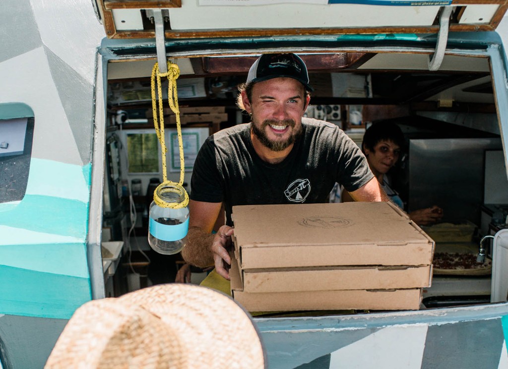 Man in hat serving pizza boxes from a food truck window.