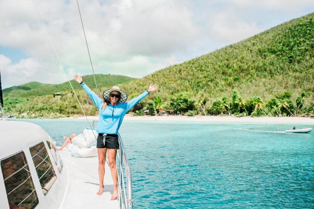 Person on a boat deck with arms raised, lush green hills and turquoise water in the background.