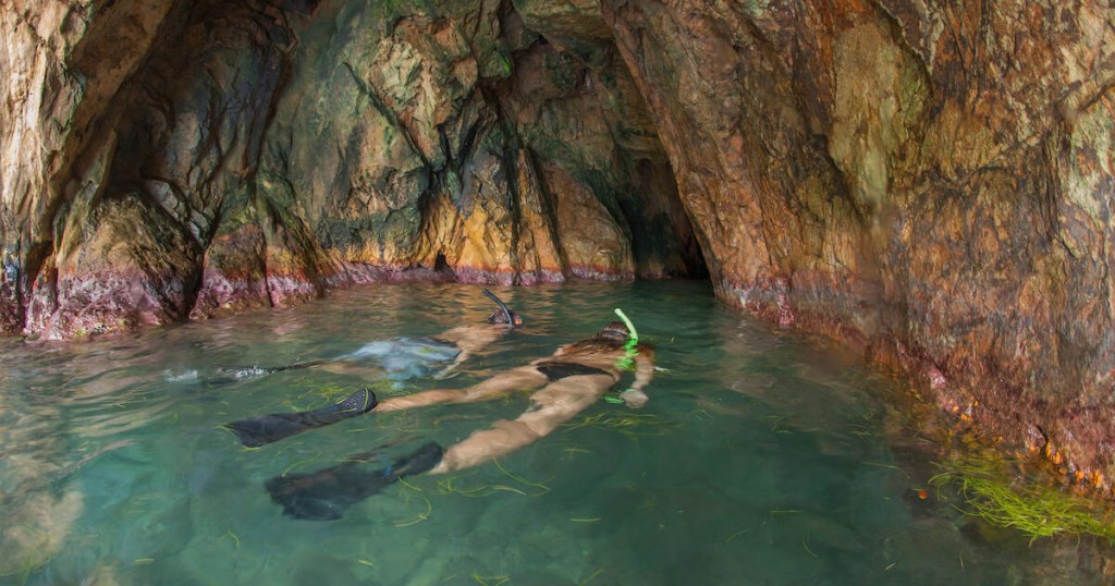 Two snorkelers swimming in a rocky cave with clear water.