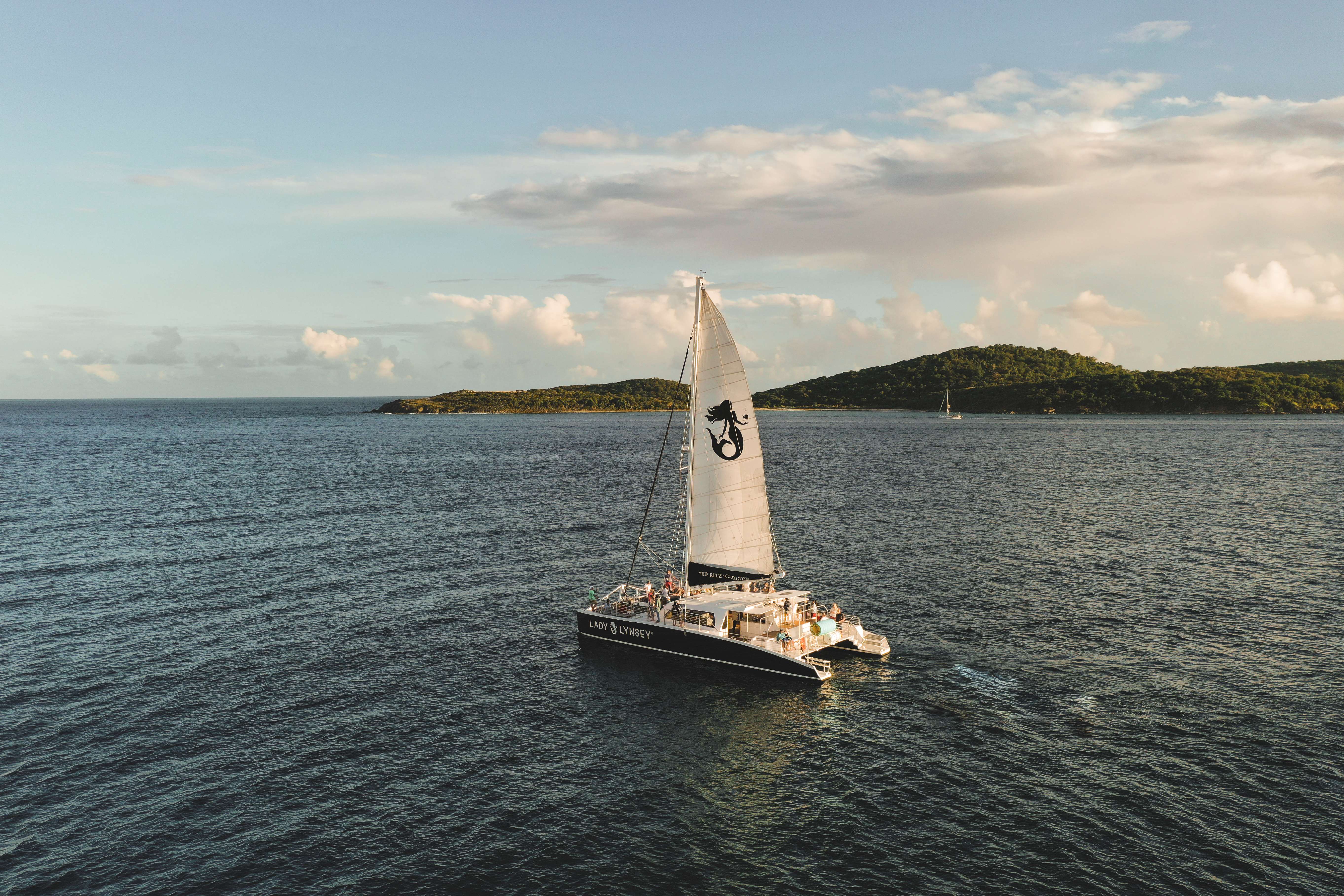 Sailing catamaran with a raised sail on calm ocean near green islands under a partly cloudy sky.