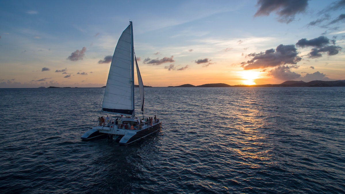 Sailing catamaran on ocean at sunset with cloudy sky.