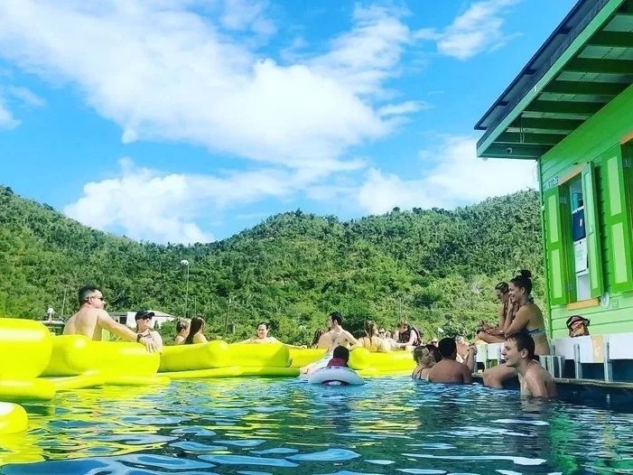 People enjoying a pool with inflatable floats near a green building, mountains in the background under a blue sky.