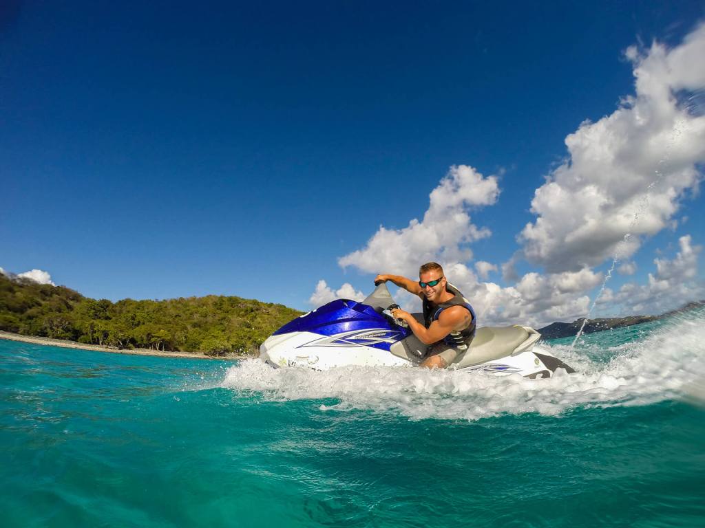 Person riding a jet ski on clear blue water near a tropical beach with a cloudy sky.