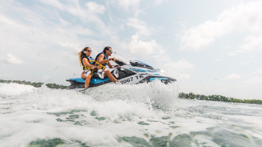 Two people riding a jet ski on a lake with splashing water and cloudy sky.