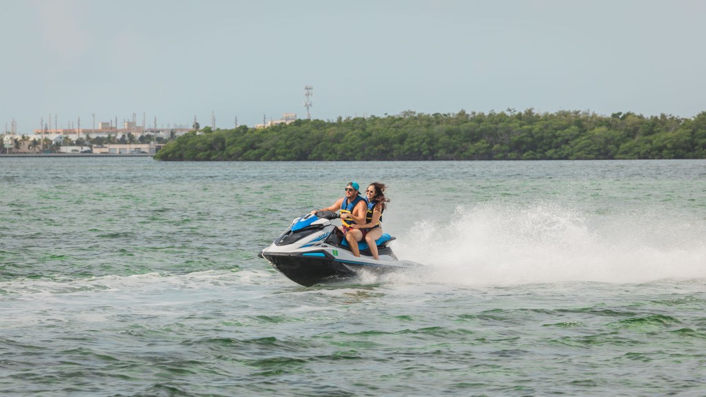 Two people riding a jet ski at high speed on a lake with forested shore in the background.