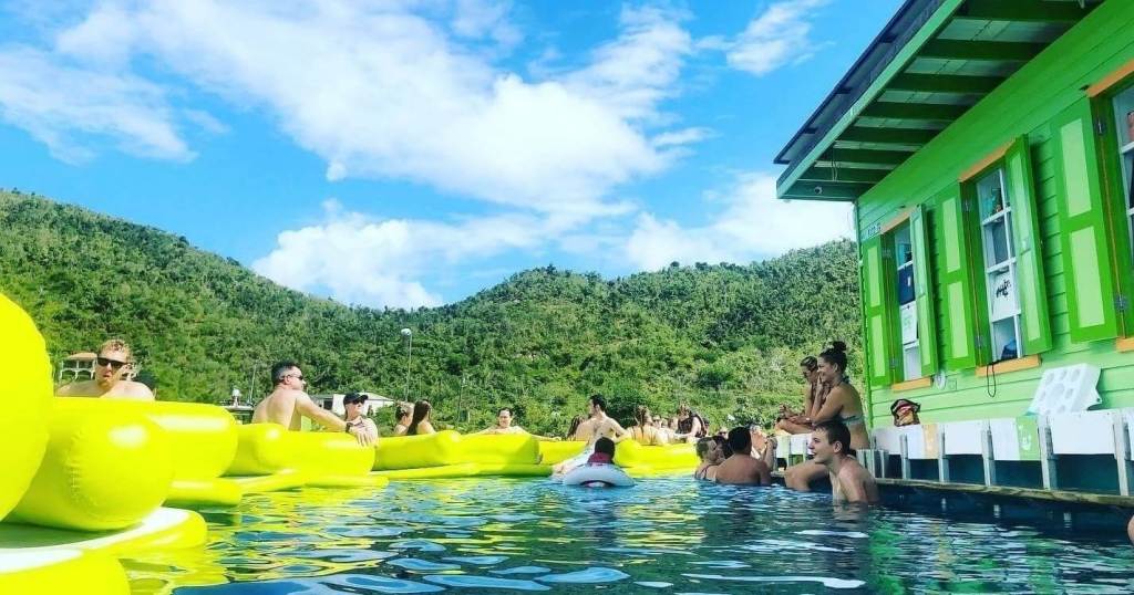 People relaxing in a pool with yellow floats near a green building and mountains.