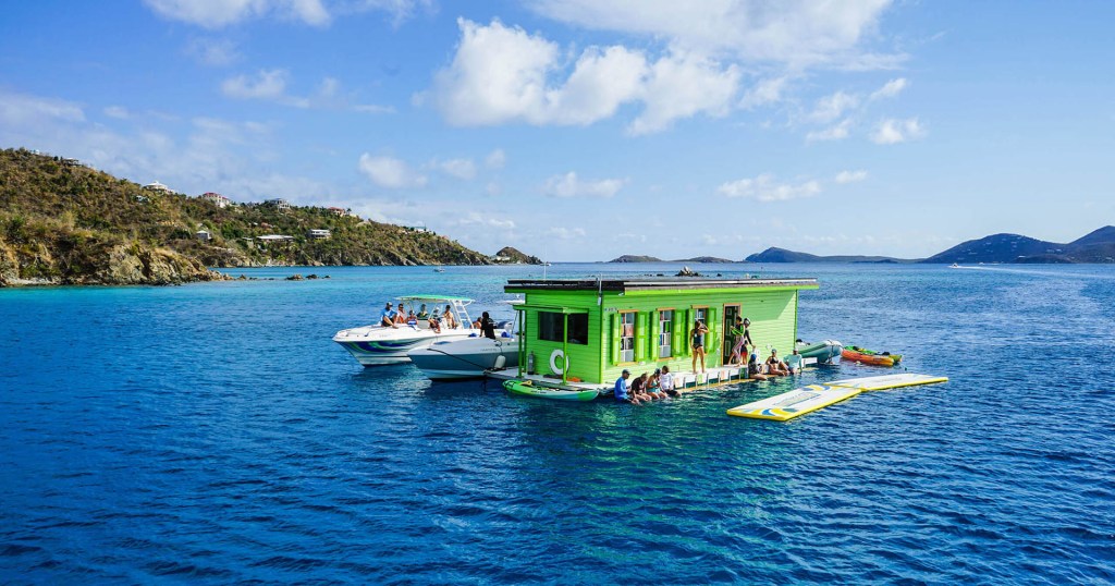 Green floating bar with people on a sunny ocean, two boats nearby, islands in the distance.