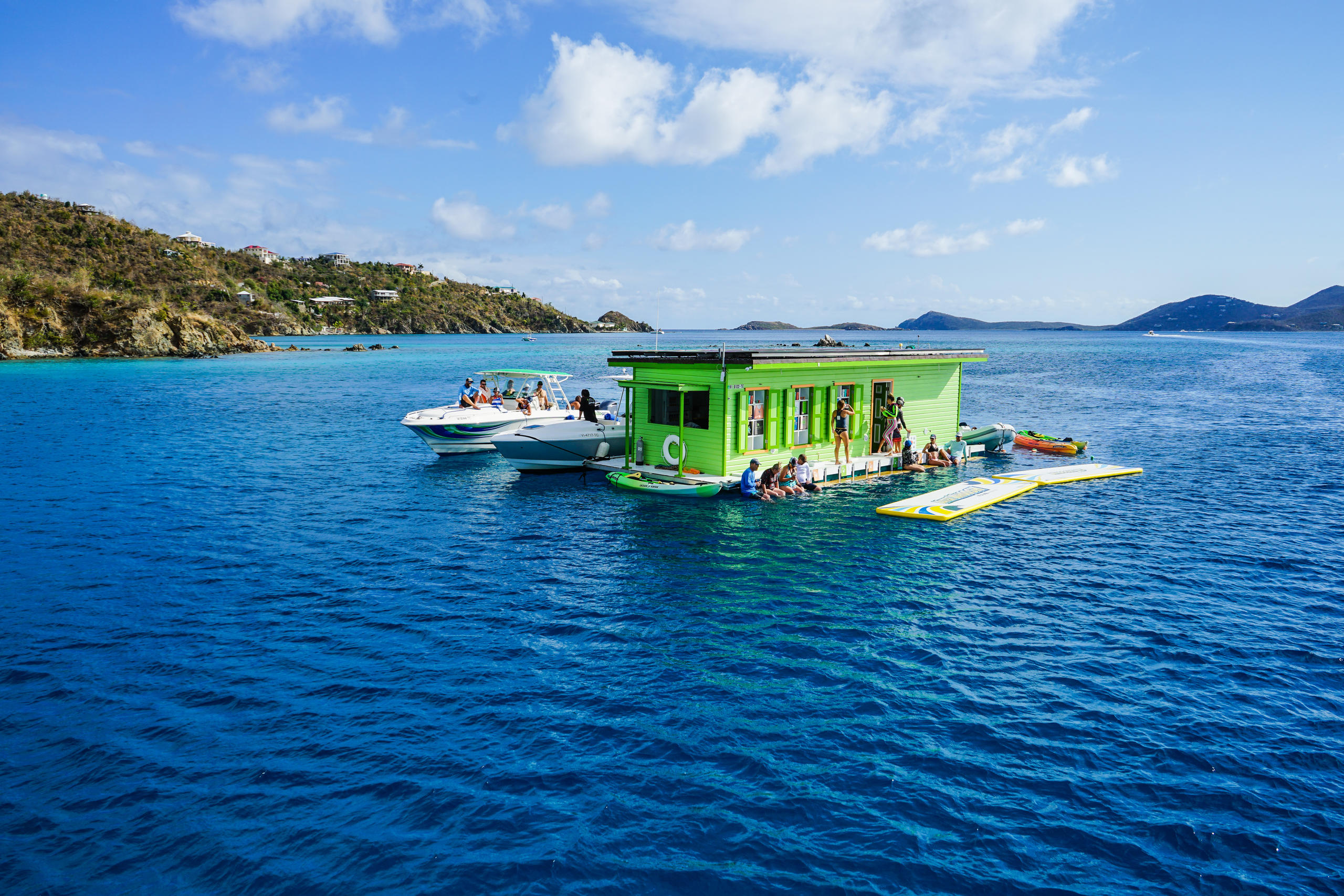 Green floating bar on blue water with surrounding boats and distant islands.
