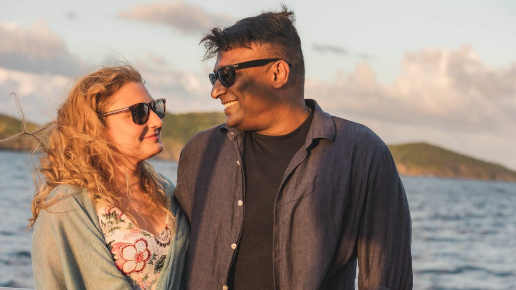 Two people smiling at each other near the sea with a scenic backdrop.