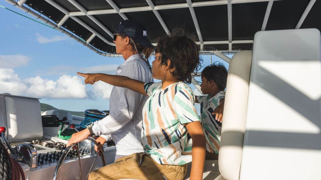 Two people on a boat, one steering and the other pointing at the horizon, under a partially cloudy sky.