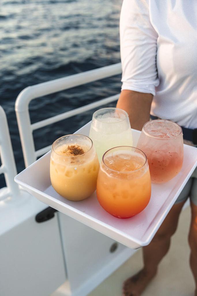 Person holding tray with four colorful drinks on a boat.