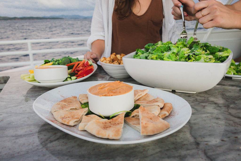 Outdoor dining with hummus, pita, salad, and vegetables on a table by the water.