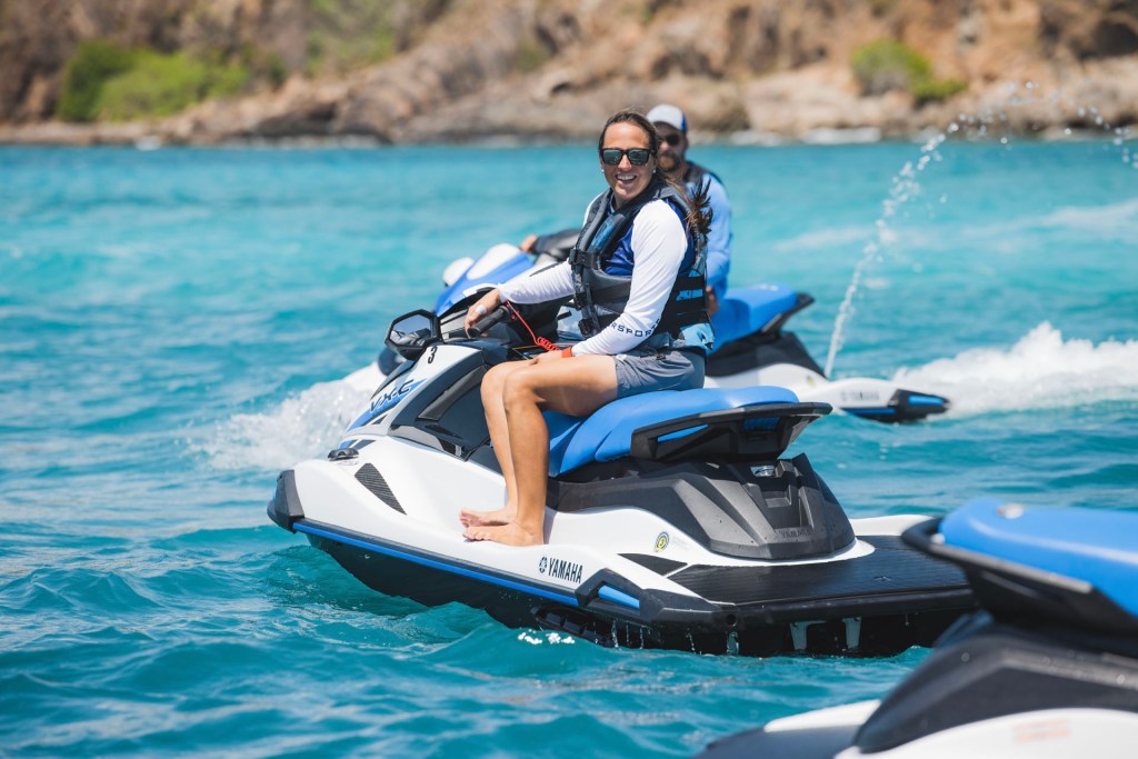 Two people riding jet skis on clear blue water near a rocky shore.