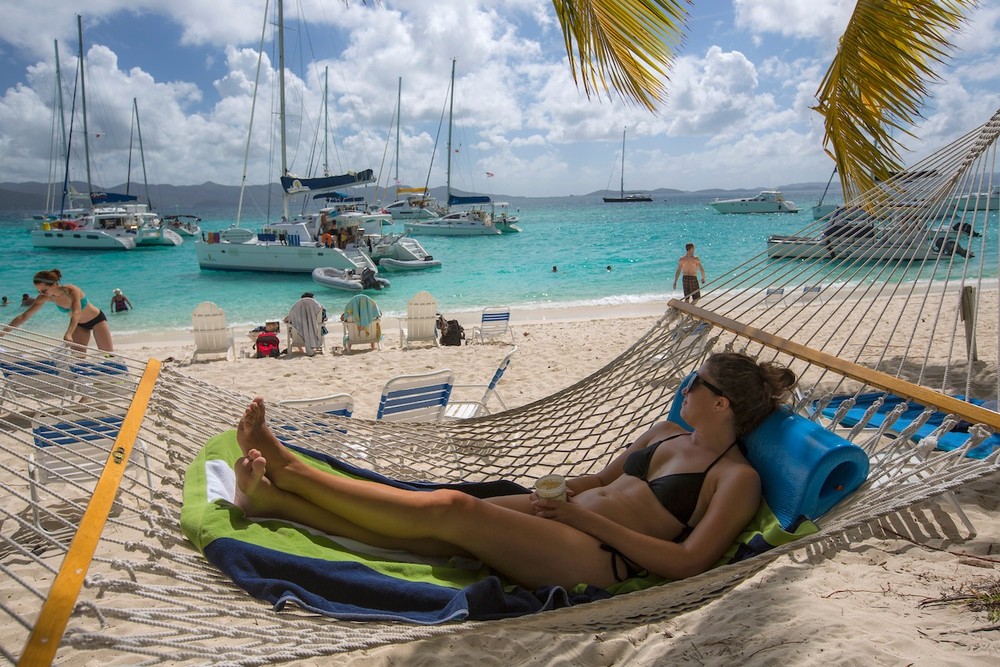 Woman in a hammock on a sunny beach, with boats on turquoise water in the background.