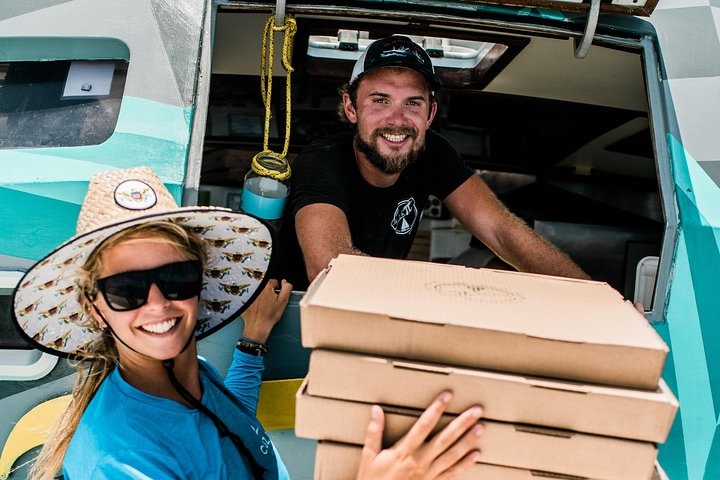 Smiling woman with hats holding pizza boxes, standing beside a man in a boat window.