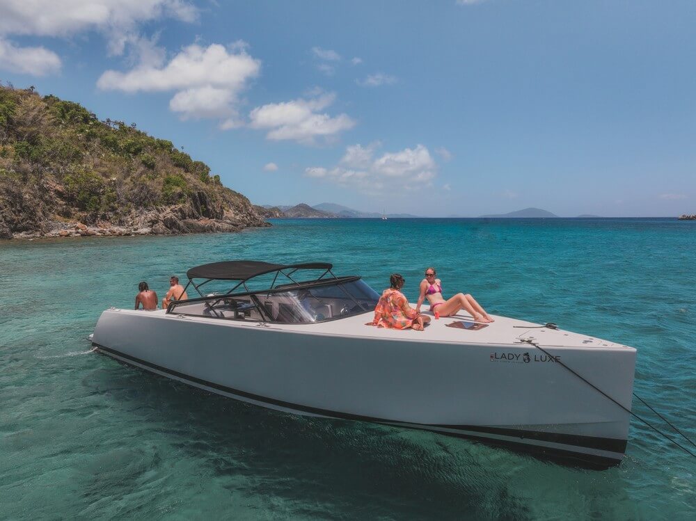White boat with people sunbathing in clear blue water near a green island under a blue sky with clouds.