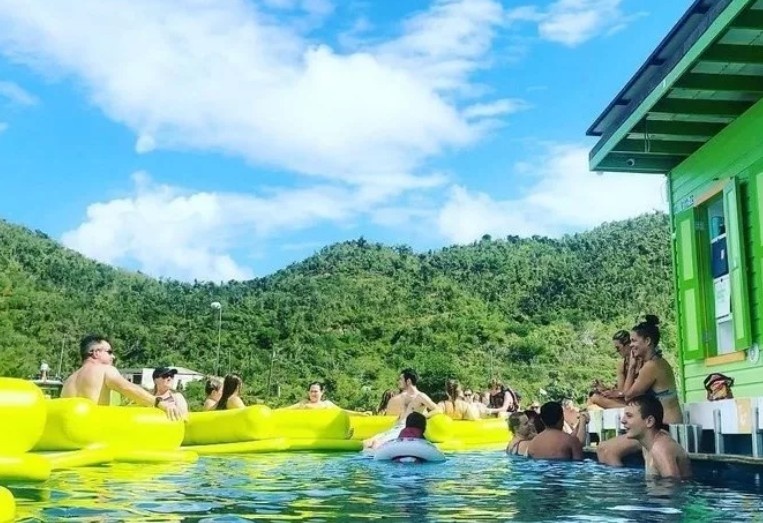 People in a pool with yellow floats by a green structure and hills in the background under a blue sky.
