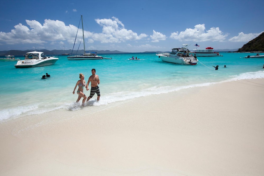 Couple walking on a sandy beach with boats on turquoise water under a clear blue sky.