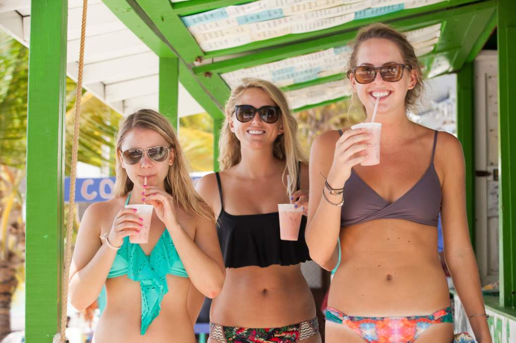 Three women in swimsuits holding drinks, standing outside a green kiosk.