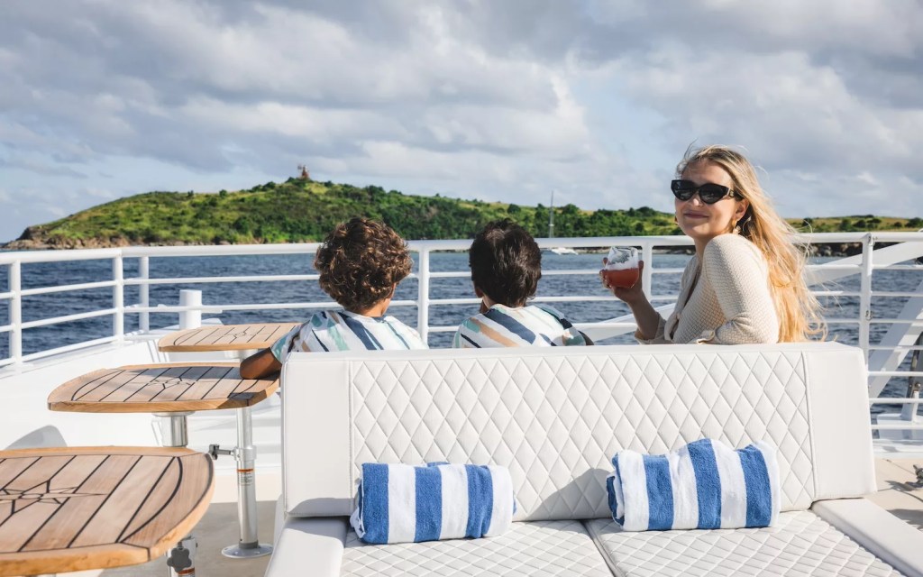 People on a boat deck with ocean and small island in background, woman holds drink, striped pillows visible.