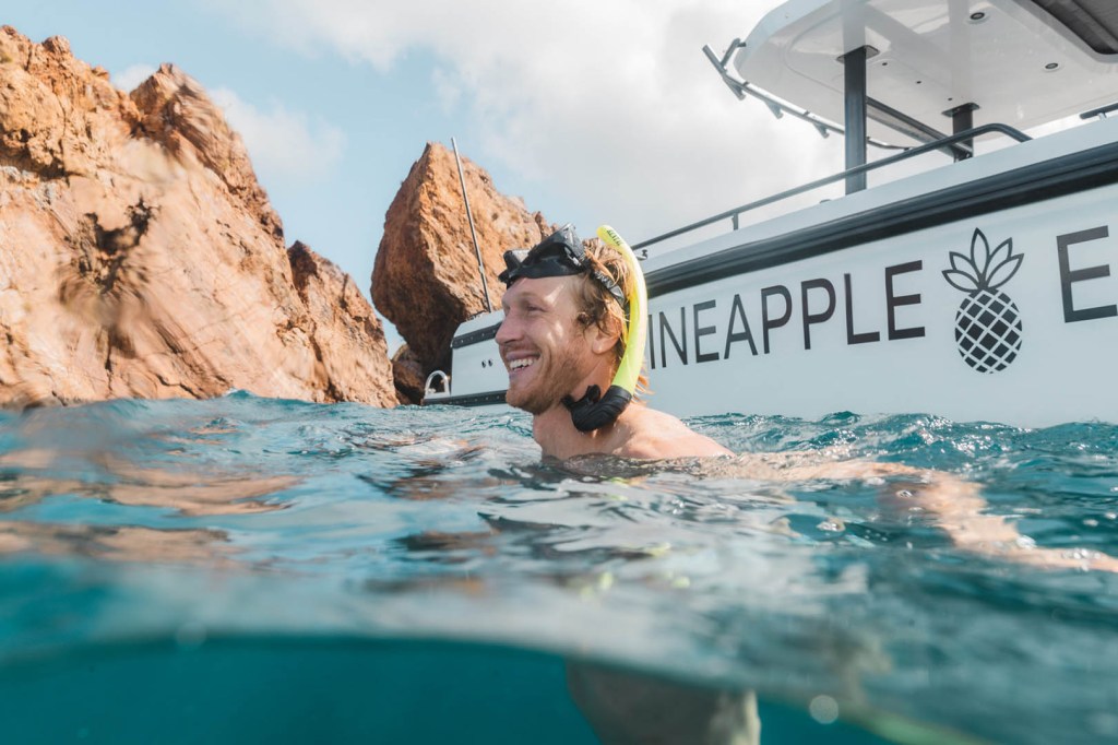 Man with snorkel smiling in water near boat and cliffs.