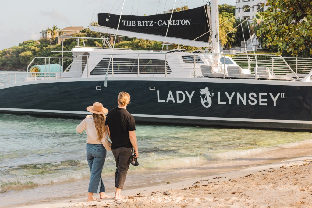 Two people on a beach looking at a boat named 'Lady Lynsey II' with 'The Ritz-Carlton' on the sail.
