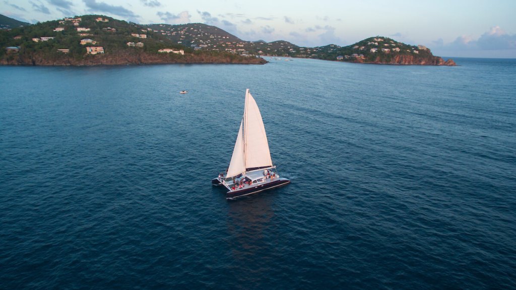 Sailboat with white sail on blue water near lush green hills during sunset.