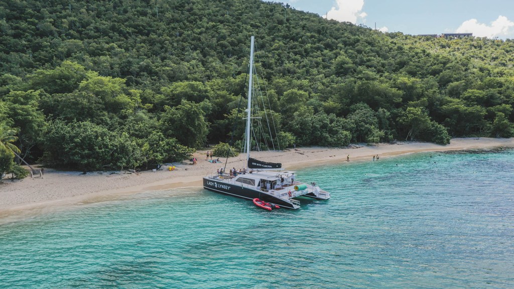 Catamaran anchored near a tropical beach with lush green hills in the background.