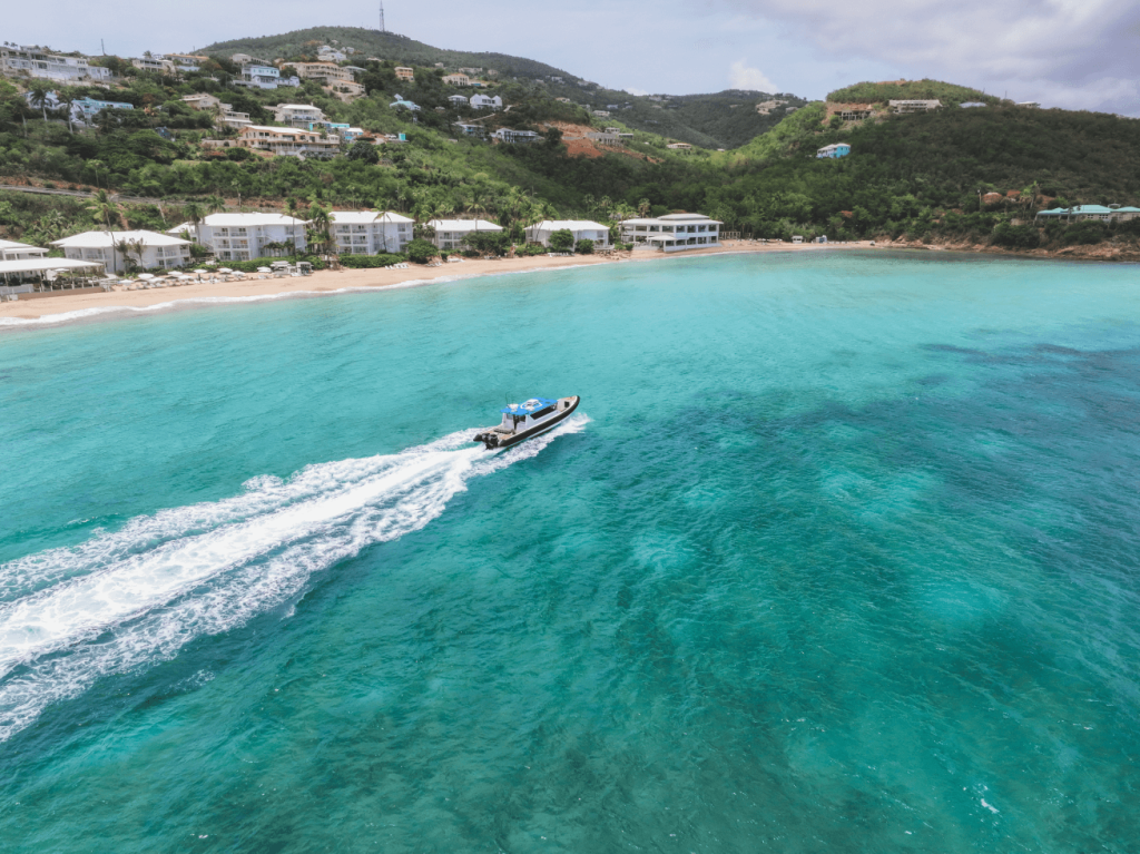 Boat speeding across turquoise water near a beach with houses and green hills in the background.