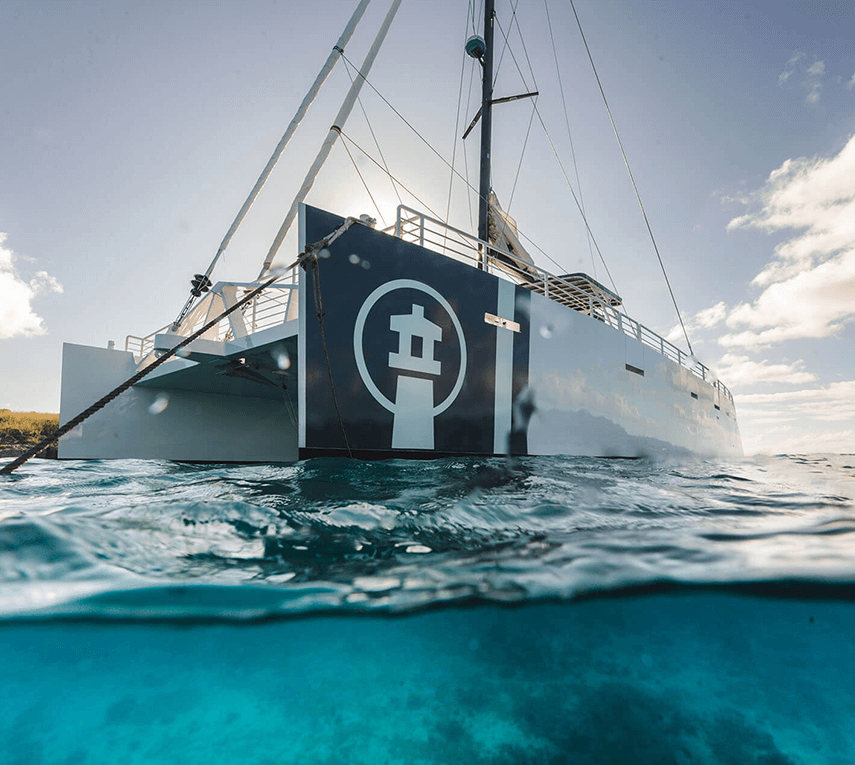 Catamaran with lighthouse logo floats on clear blue water with cloudy sky.