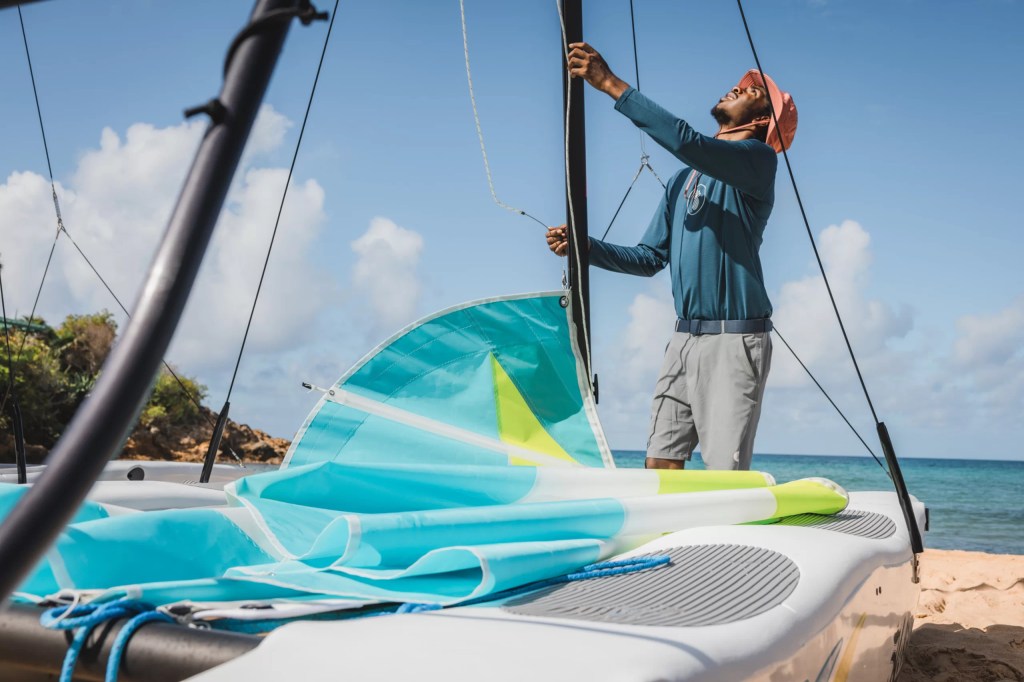 Person preparing sails on a catamaran at a sandy beach with blue sky and sea.