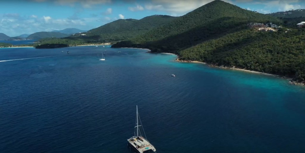 Sailboats on water near forested mountain coastline under blue sky.