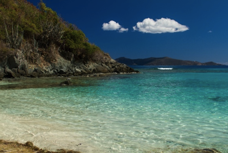 Tropical beach with clear turquoise water, rocky coast, and a single cloud in a deep blue sky.