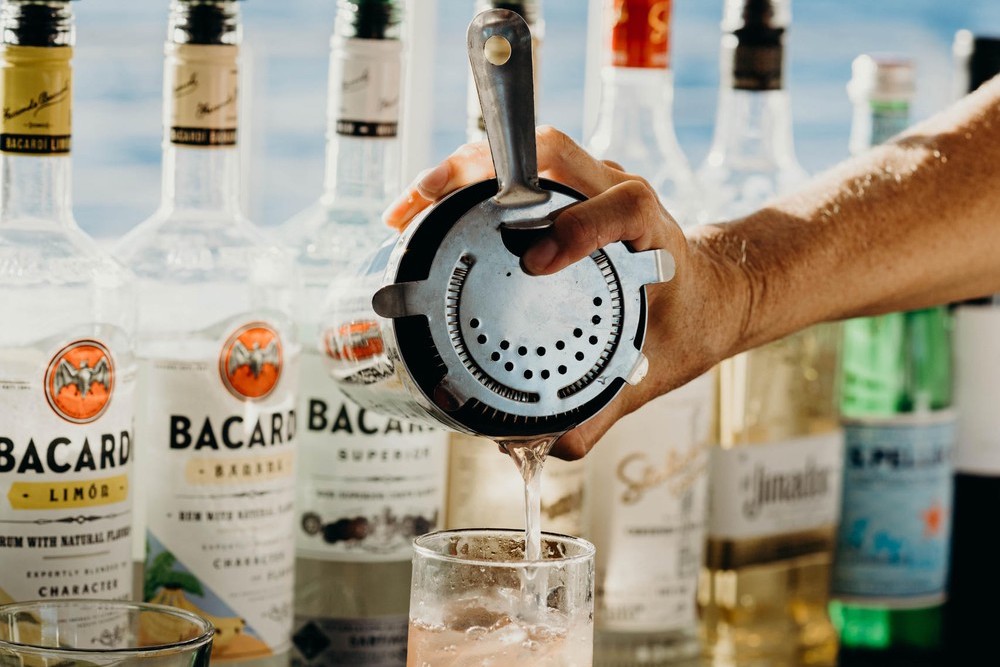 Hand pouring drink through strainer into glass, Bacardi bottles in background.