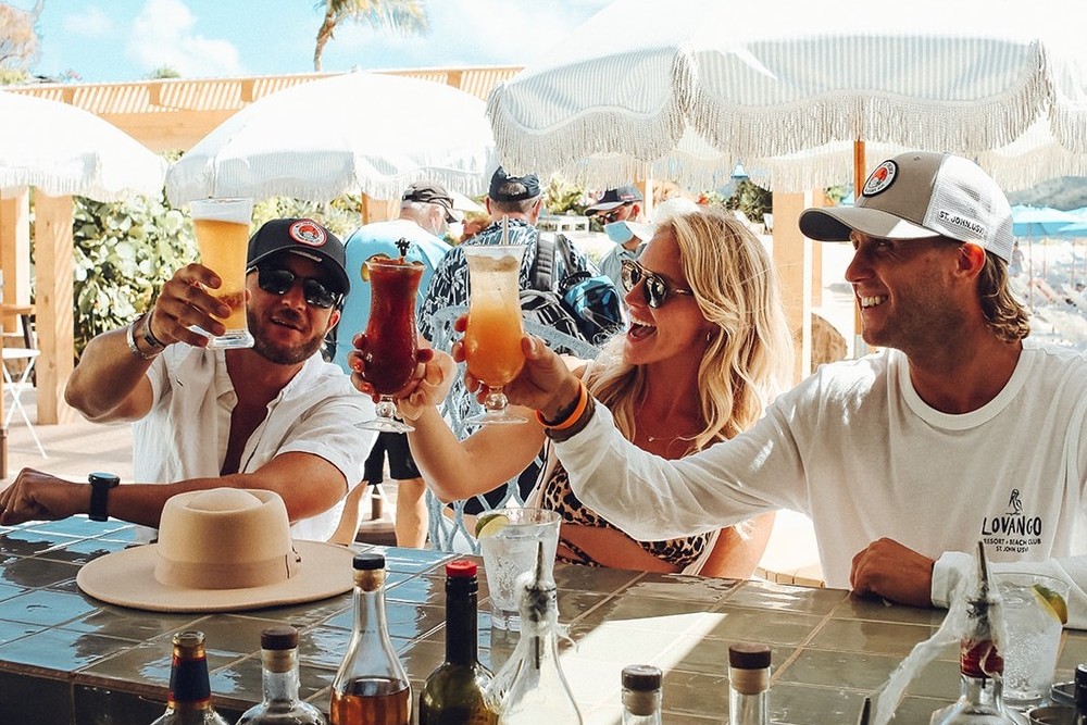 Three people at a bar clinking colorful cocktails under sun umbrellas.