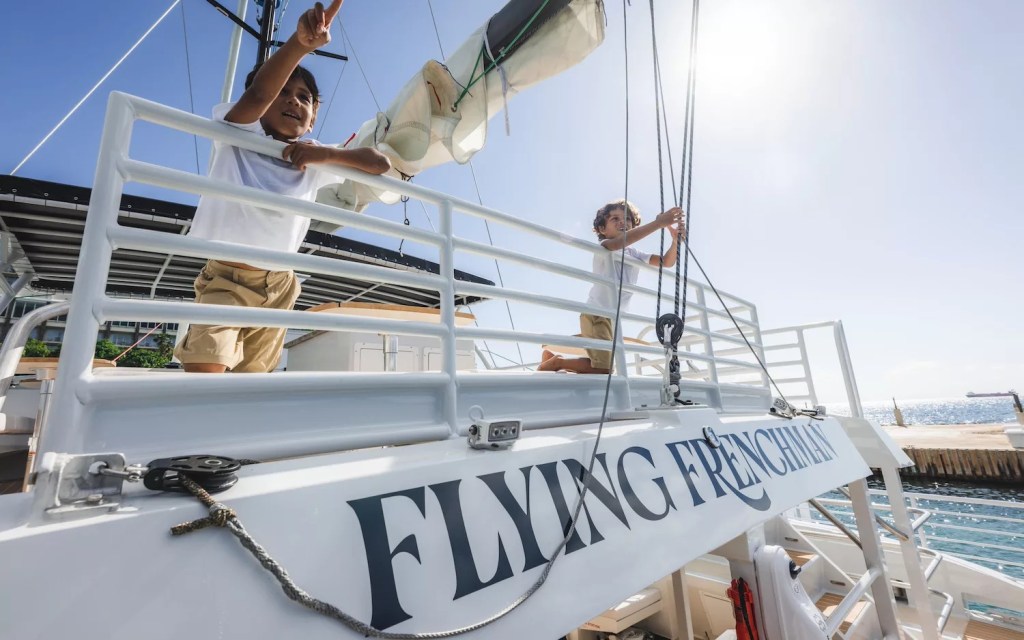 Two people on a sailboat named 'Flying Frenchman,' with ropes and sails in the background.