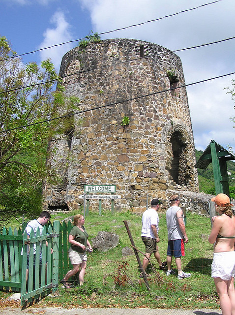 Tourists walk near a historic stone tower with a 'Welcome' sign and green fence.