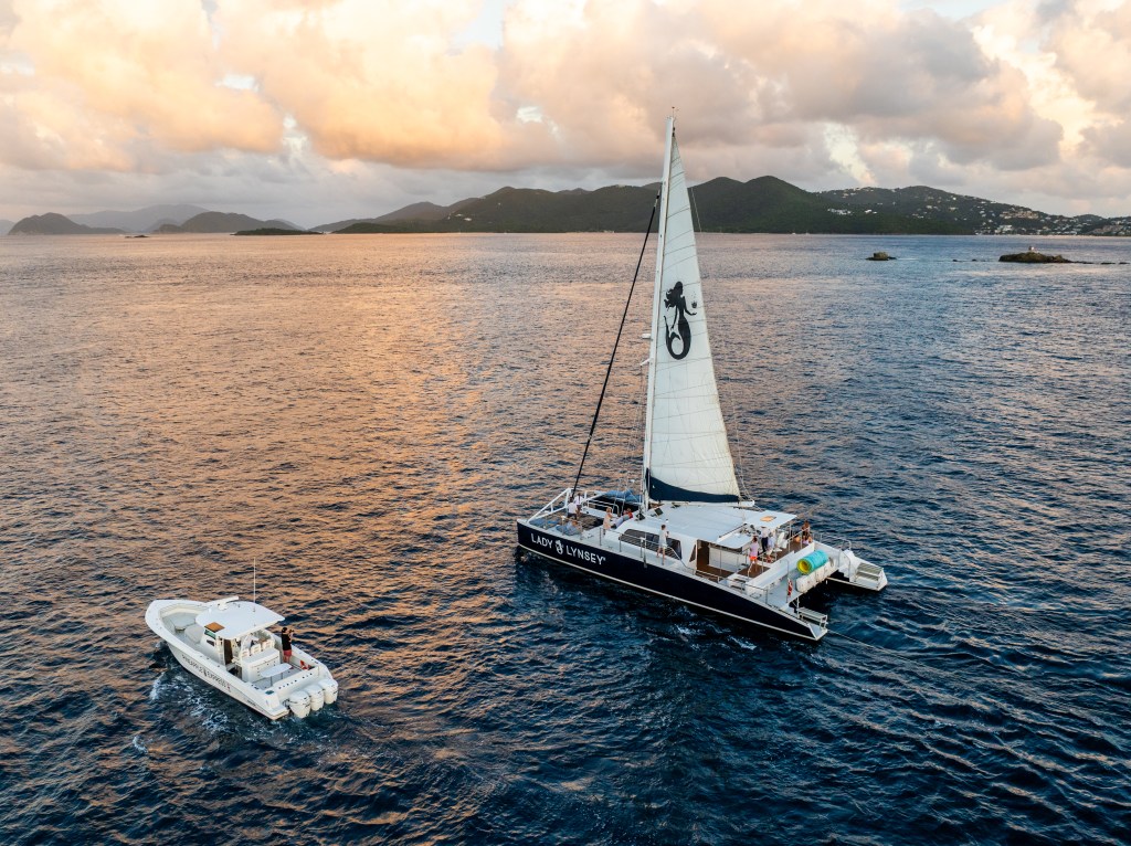 Sailboat and motorboat on calm sea with islands and cloudy sky at sunset.