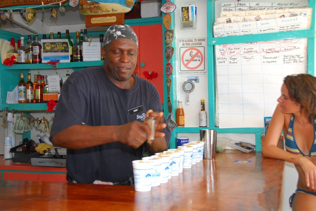 Man behind bar preparing drinks, with woman seated at counter watching.