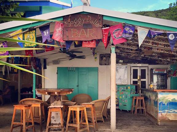 Outdoor tropical bar with stools, colorful flags, and rustic decor under a canopy.