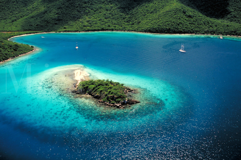 Small island with lush greenery in a turquoise bay, surrounded by boats and green hills.
