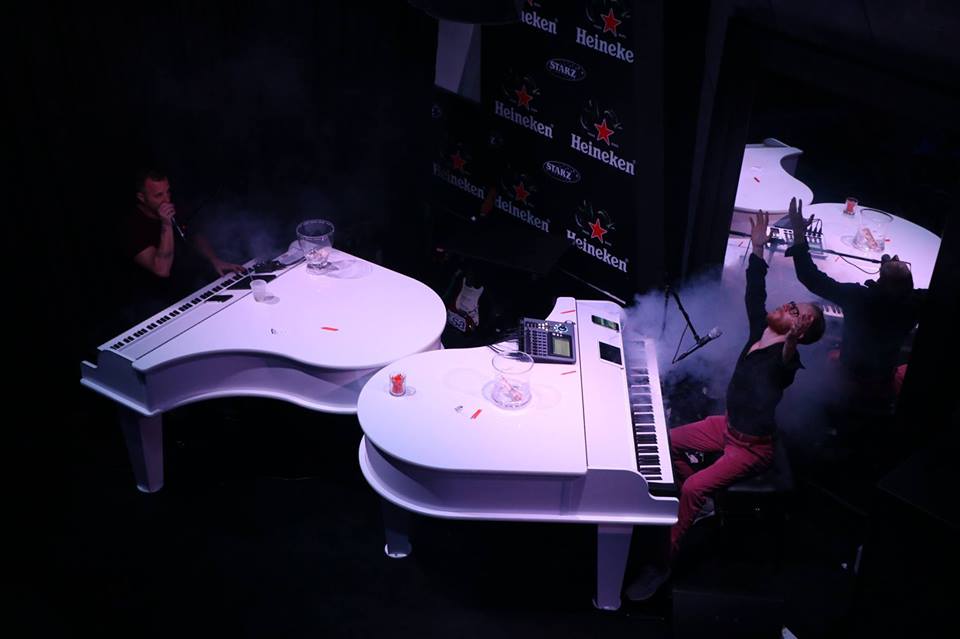 Two musicians playing white pianos with dramatic stage lighting.