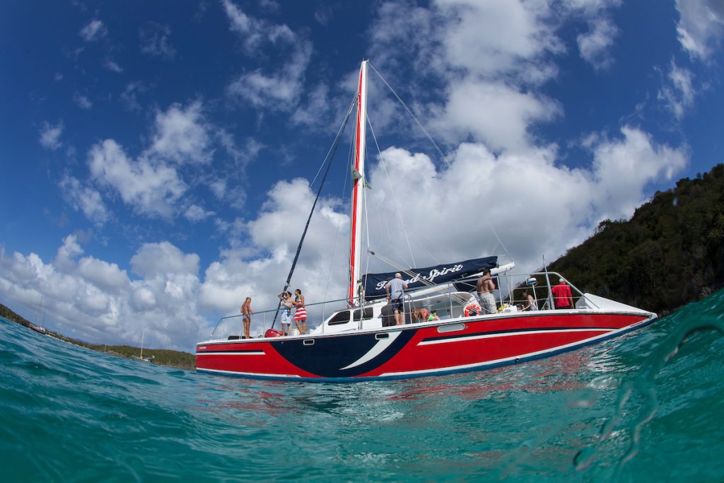 Red sailboat with people on deck sailing in clear waters under a partly cloudy sky.
