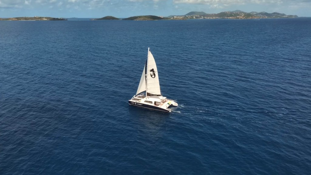 Sailboat with number 3 sails on blue ocean near distant hilly islands under a partly cloudy sky.