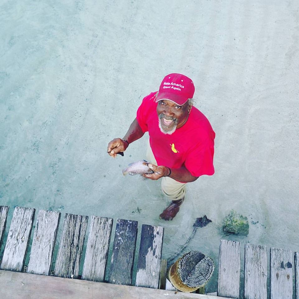 Man in red shirt and hat smiling, holding a fish near a wooden pier.