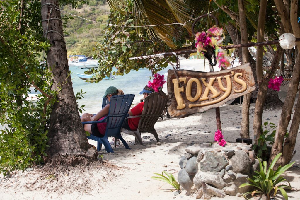 Beach scene with Foxy's sign, two people on chairs, and tropical plants.